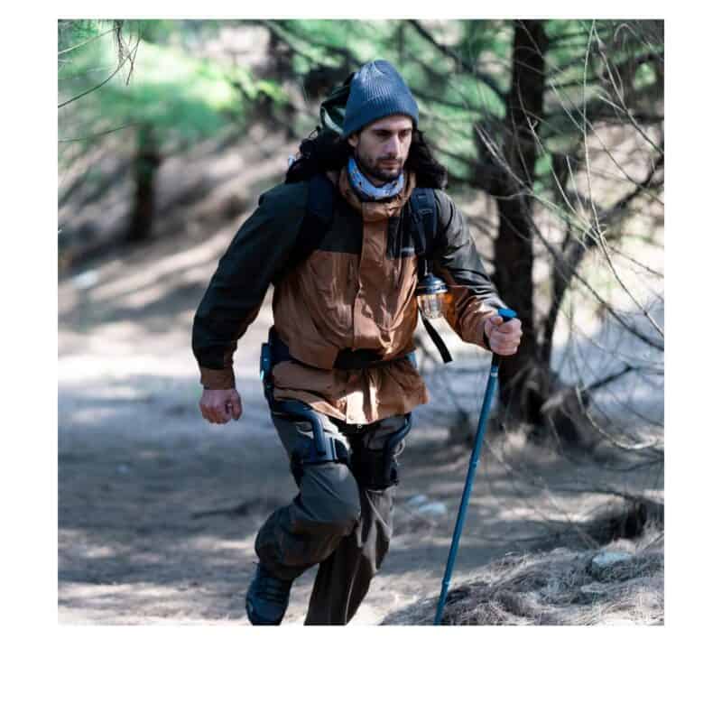 Escursionista in salita su un sentiero nel bosco con l'esoscheletro per le anche DNSYS X1 indossato in vita e i bastoncini da trekking